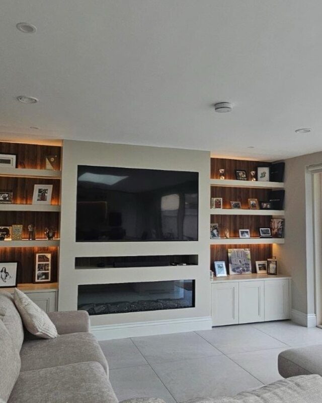 A modern built-in media wall unit designed to be the focal point of a living room. Here’s a clear breakdown of its features and design elements:
Central TV & Fireplace Section

A flat-screen Tv is mounted centrally within a clean, recessed section.
Beneath the TV is a linear electric fireplace, giving the wall a sleek, contemporary look.
The TV and fireplace are framed symmetrically.

On both sides of the central section, there are custom built-in shelves that run from floor to ceiling.
Each shelf is backlit with warm LED lighting, creating an inviting glow and highlighting the items displayed.
The back panels of the shelves have a wood finish, adding texture and warmth to the otherwise minimalist space.

Lower Cabinets
The lower portion on both sides features closed cabinetry with subtle vertical paneling and hand painted .
These provide hidden storage, helping keep the space tidy by concealing electronics, cables, or miscellaneous items.

 The entire area creates a clean, modern, and cozy entertainment area. #mediawall #tvunit #bespokefurniture